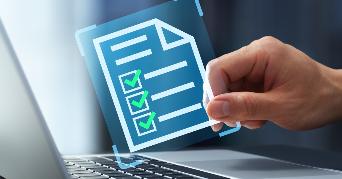 A hand holds a digital checklist with green check marks above a laptop keyboard. The background is blurry.