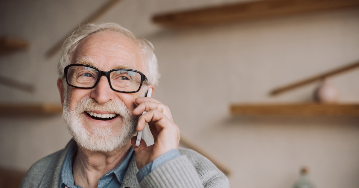 A smiling older man with glasses and a white beard holds a smartphone beside his face as he stands near wooden shelves.
