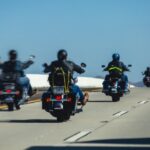 A group of motorcyclists rides across multiple lanes on a highway under a clear blue sky with distant hills.