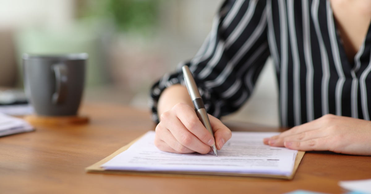 A person in a black striped shirt writes on a paper document at a wooden desk with a gray mug nearby.