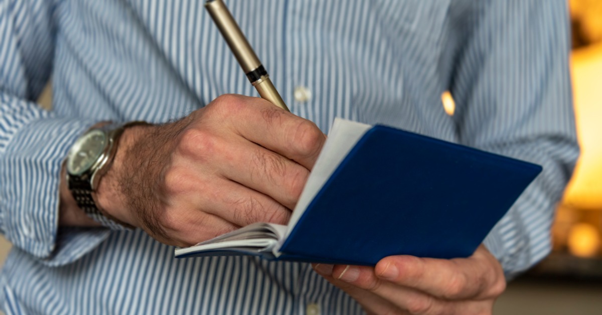 Close-up of a person's hands writing in a small blue checkbook with a hold pen. The person wears a blue and white shirt.