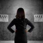 A woman in a dark suit stands with her hands on her hips and looks at two sketched arrows on a textured gray wall.
